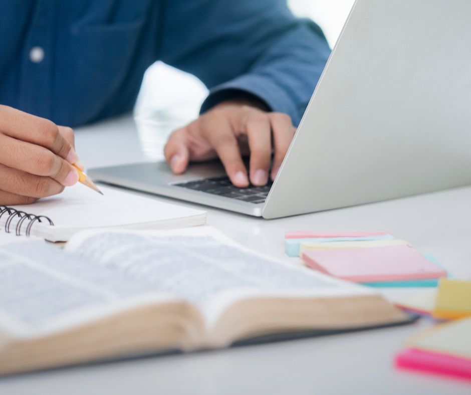 Person studying with an open book, laptop, and notes on a white desk.