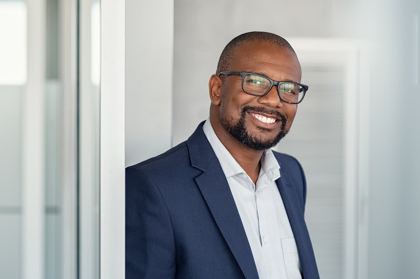 Smiling African-American man in a professional blue suit with a white shirt, wearing glasses, standing in a modern office environment.