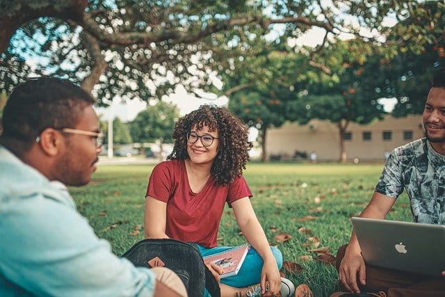 Students in casual attire having a discussion on a grassy campus, with one holding a tablet and another using a laptop, conveying a relaxed outdoor study session.