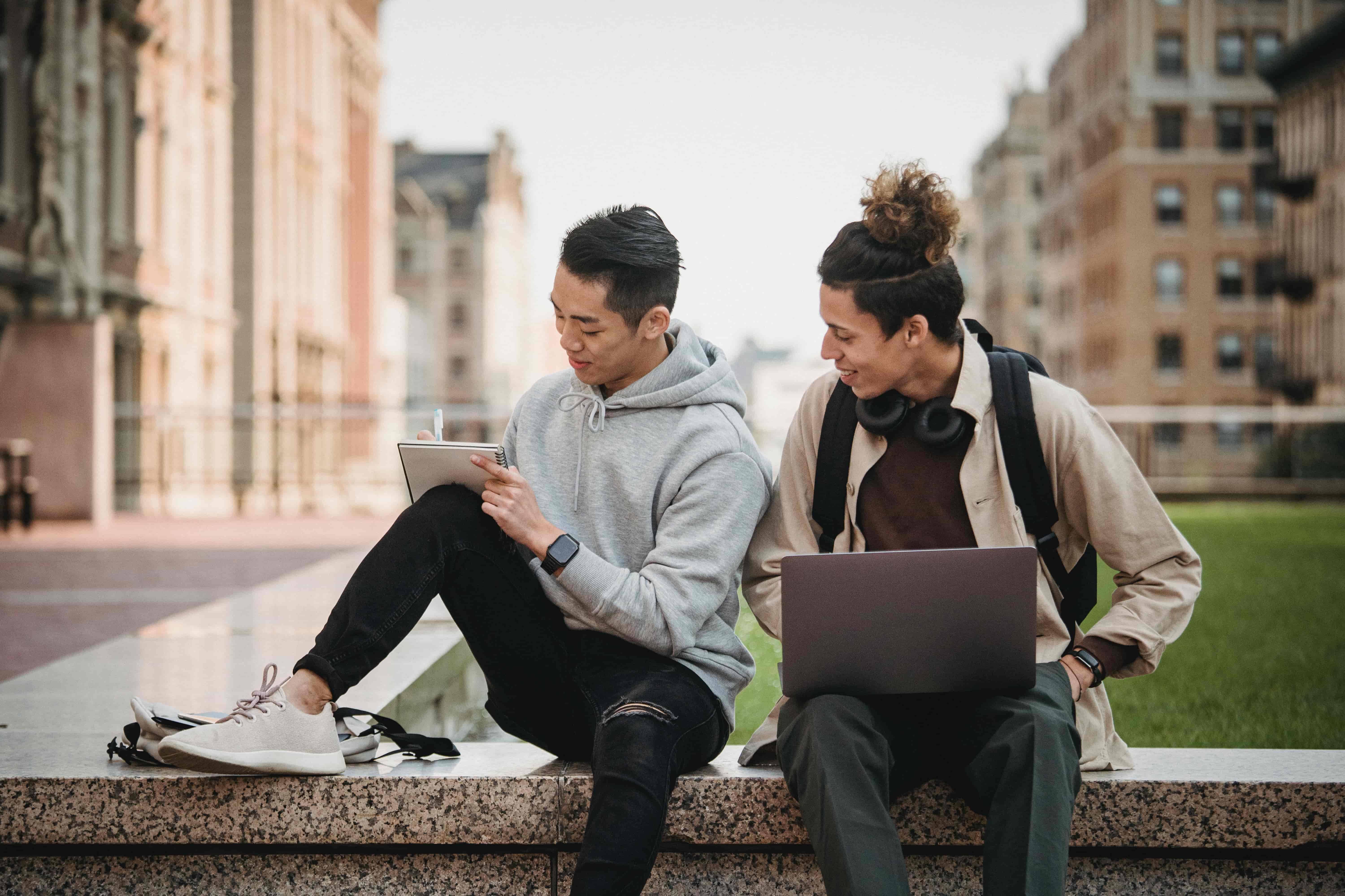 Two students engaged in studies; one is writing in a notebook with a pen and the other with a laptop.