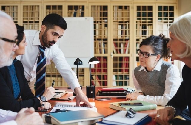 Institutional team meeting with a man gesturing towards documents and discussing with colleagues around a conference table.