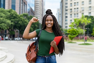 Joyful Nigerian student with braided hair, holding a red notebook and raising a fist.