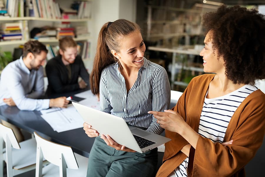Two female coworkers smiling at each other. One holds a laptop