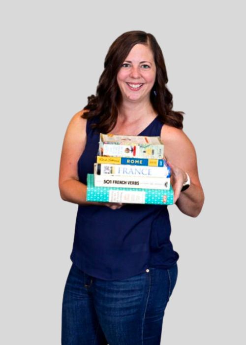 Amy Kawa beams holding a stack of travel books including guides on Rome and France, and '501 French Verbs'.