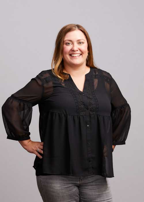 Amy Ullrich smiles confidently, hands on hips, in a sheer-sleeved black blouse and gray jeans.