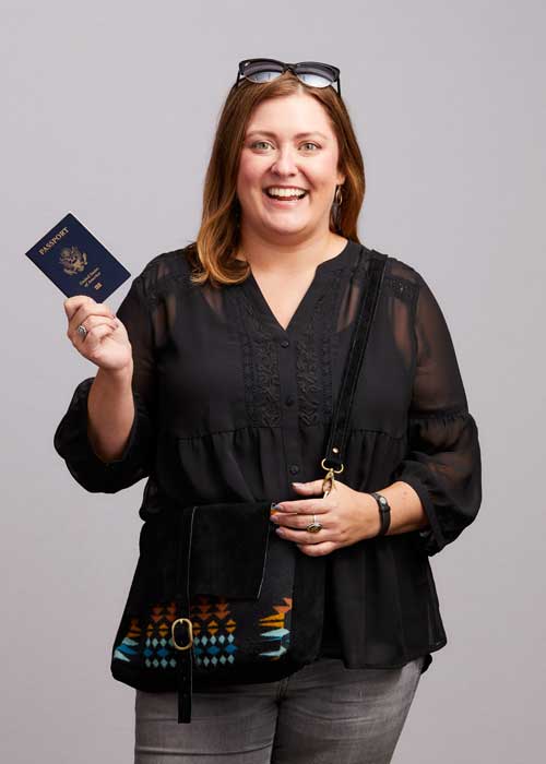 Amy Ullrich holds up her passport, ready for travel, in a chic black blouse with a patterned purse.