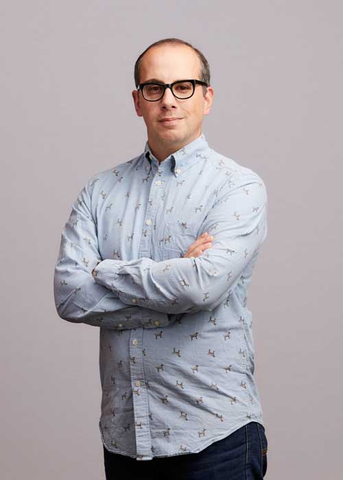 Dan Agacki in a playful, animal-patterned shirt, confidently posing with arms crossed.