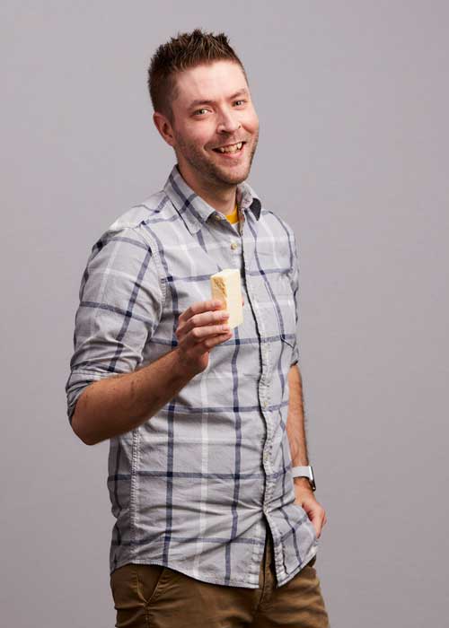 David Jarvela shares a grin, holding a snack, in a casual gray plaid shirt.