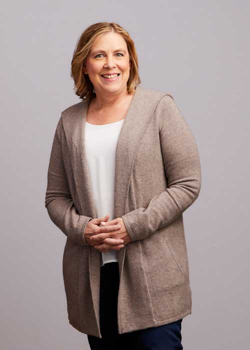 Heidi Ames posing with a warm smile, hands clasped in front of her, wearing a beige cardigan and white top.