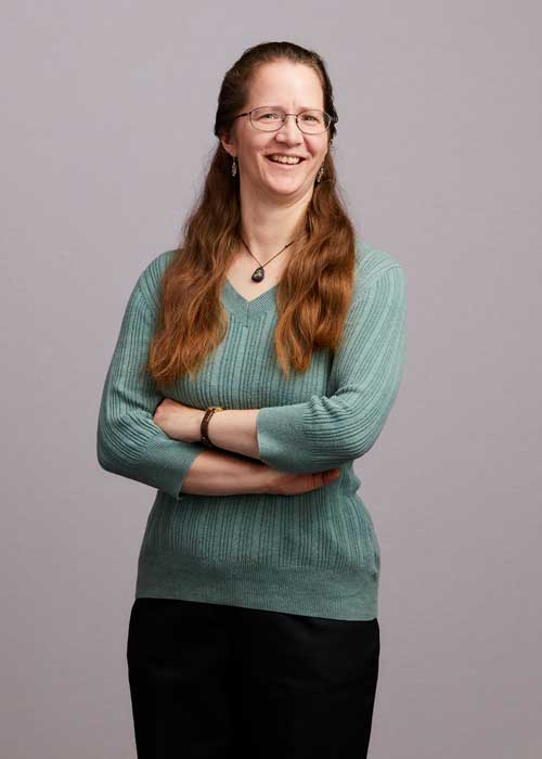 Karen Krug, smiling contentedly with her arms crossed, is wearing a cozy teal sweater paired with a black skirt.