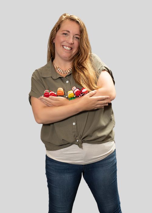 Kelsey Ward laughs while holding a set of teeth toys in her crossed arms, wearing a khaki green button-up shirt.