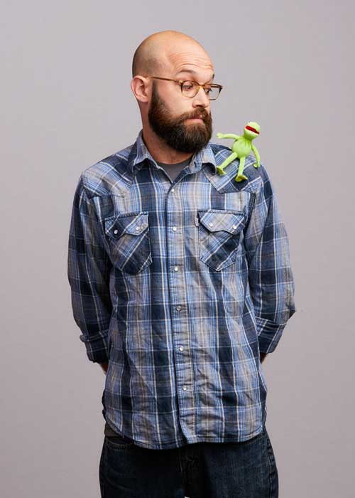 Kevin DeMars with a whimsical expression, a Kermit the Frog toy perched on his shoulder, wearing a blue plaid shirt.