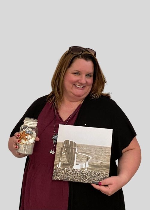 Kimberly Hajec smiles brightly, holding a mason jar with seashells and a coastal-themed photo canvas, wearing a black cardigan.