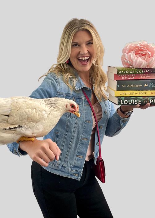 Lauren Usma grinning while holding a stack of books, with a hen perched on her arm, dressed in a denim jacket.