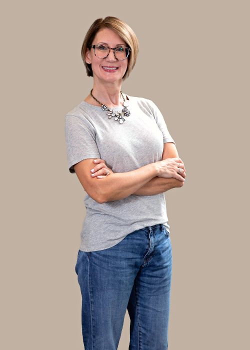 Leigh Lane Peine with arms crossed, wearing glasses, a silver necklace, and a casual grey top.