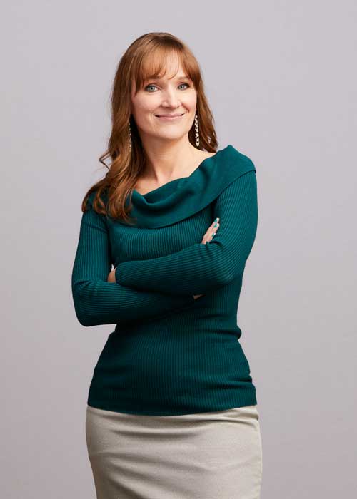 Majka Drewitz with a charming smile, arms folded, wearing a teal sweater and a cream-colored skirt.
