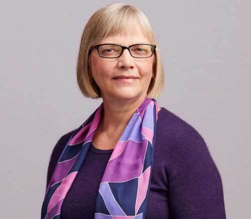 Margit Schatzman, smiling with glasses, purple sweater, and patterned scarf.