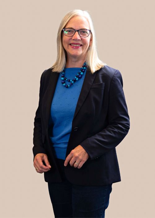 Margit Schatzman in professional attire, with a tailored black blazer over a bright blue sweater and a bead necklace.