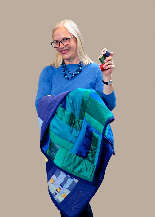 Margit Schatzman holding a vibrant blue and green quilt and a spool of thread, smiling in a coordinating blue sweater.