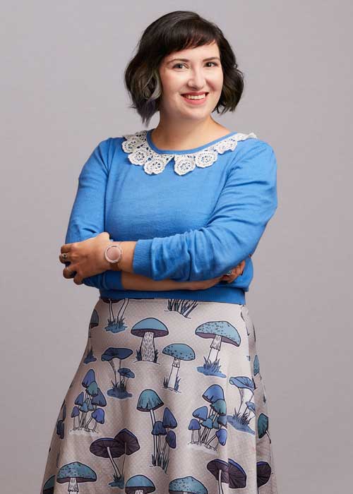 Mary Dumke standing confidently with arms crossed, dressed in a blue sweater with a unique mushroom-print skirt.