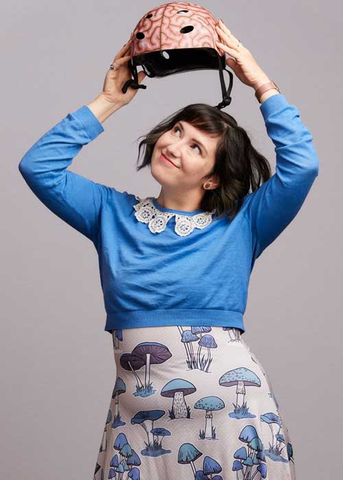 Mary Dumke playfully balancing a pink bicycle helmet on her head, wearing a bright blue sweater with a lace collar.