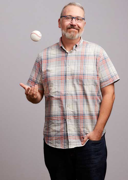 Matthew Holochwost casually tossing a baseball up in the air, wearing a short-sleeved plaid shirt with a smile.