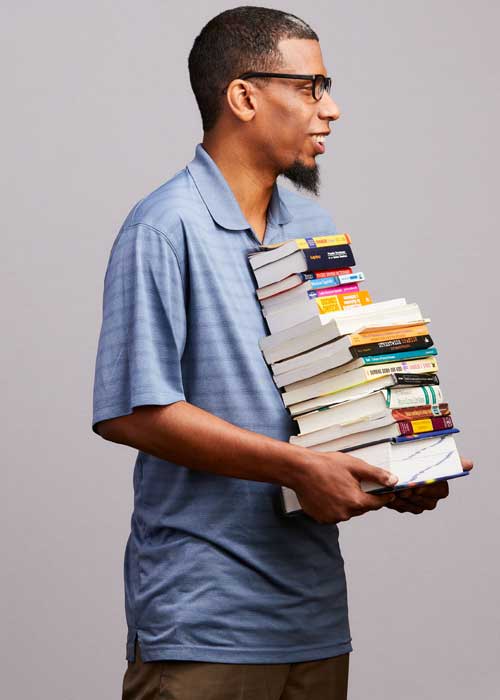 Michael Payne carrying a hefty stack of books, dressed casually in a blue polo shirt.