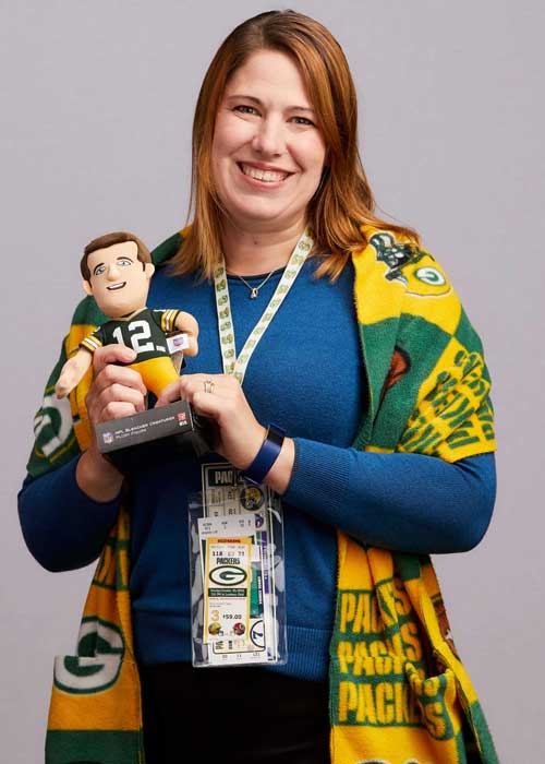 Sheree Yates proudly holding a Green Bay Packers-themed blanket and bobblehead, with a bright blue sweater.