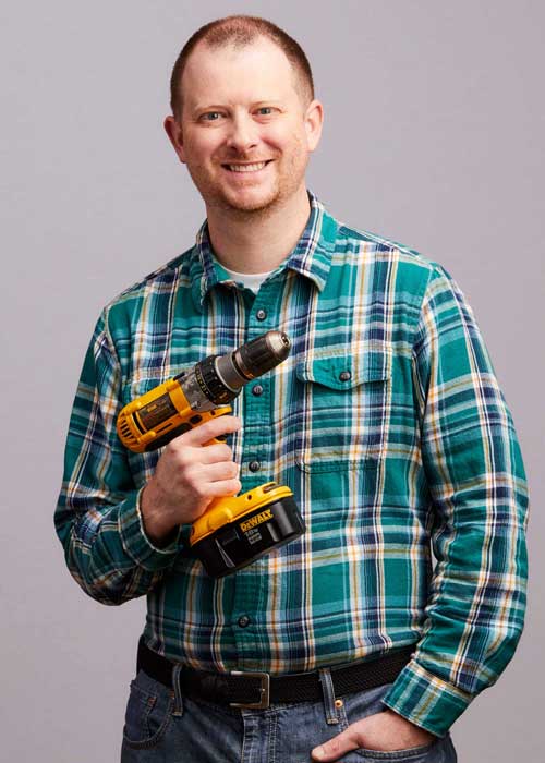 Timothy Kell smiling with confidence, holding a yellow and black DeWalt drill, wearing a green plaid shirt.