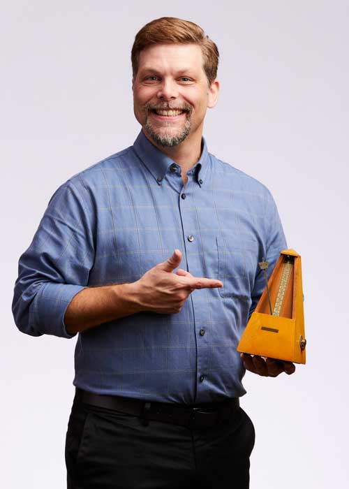 Thomas Robinson playfully pointing at a vintage yellow metronome, wearing a blue checked shirt.