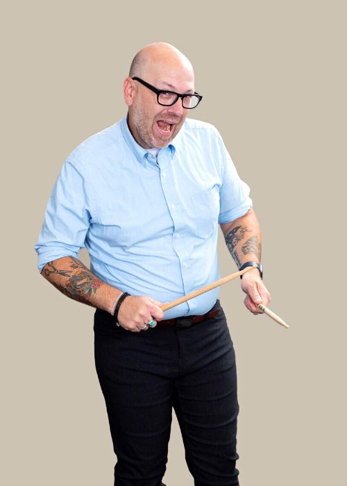 Zachary Holochwost in a playful pose, holding drumsticks and making an excited face, showcasing his tattooed arms.